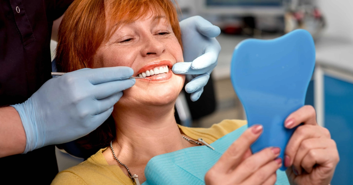 Woman smiling during a dental checkup, looking in a mirror while the dentist examines her teeth, showcasing healthy results after Dental Implants in Chandler, AZ.
